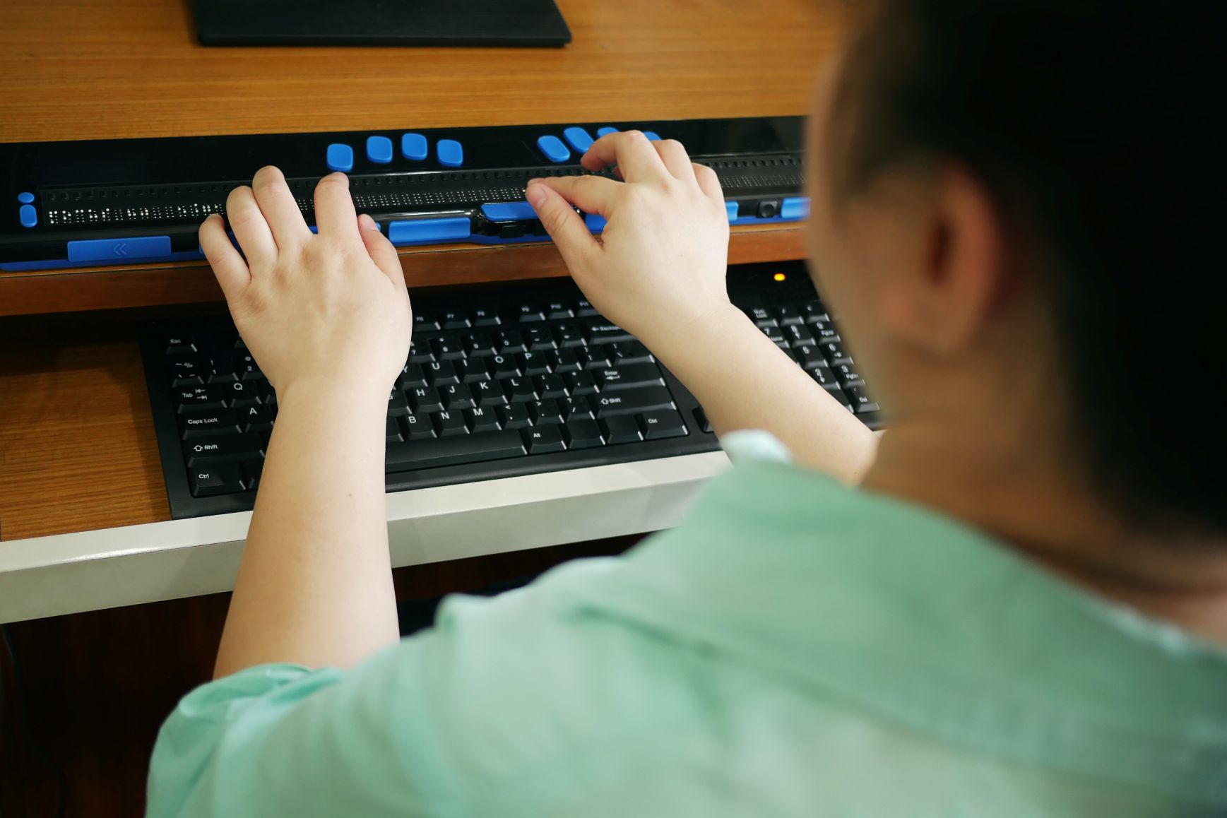 Rear view of blind female student using braille display assistive technology with computer.